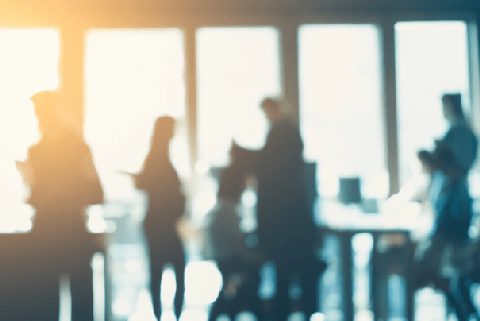 Verschwommene Silhouetten von vier Menschen, die in einem hellen Büro mit großen Fenstern stehen und sich unterhalten.